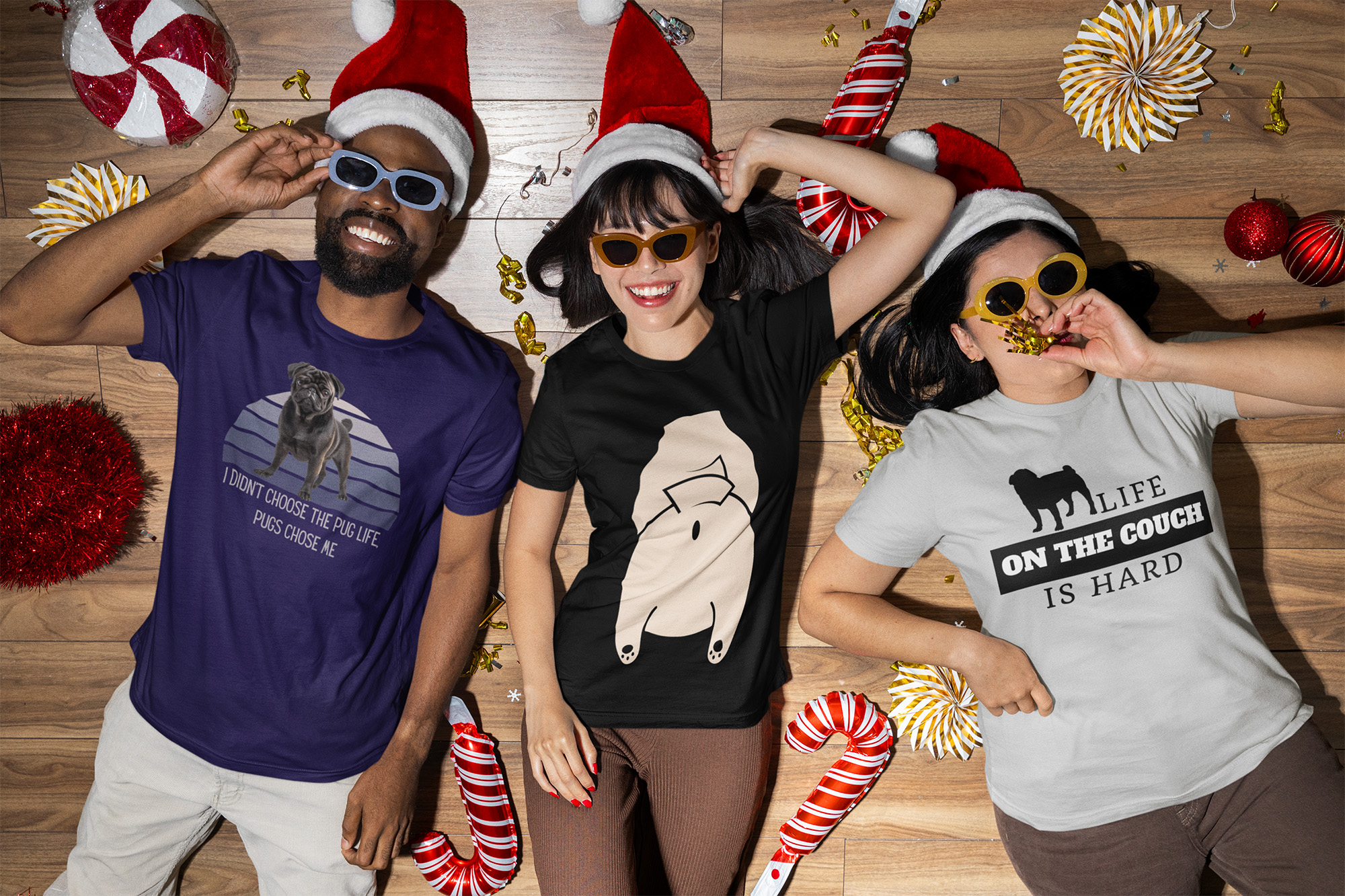 Three people laying on a hardwood floor wearing Christmas hats. Large stuffed candy canes are scattered around them. All three are wearing pug life t-shirts.