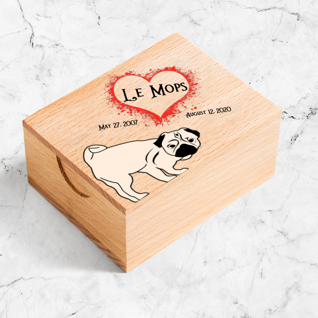 Wooden box on marble counter. On the top of the box is an illustration of a pug with the words Le Mops and the dates May 27, 2007 to August 11, 2020. Le Mops is framed in a heart.